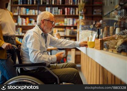 Disabled grandfather in wheelchair and granddaughter at the counter, disability, cafeteria interior on background. Handicapped older man and young female guardian, paralyzed people in public places. Handicapped older man and young female guardian