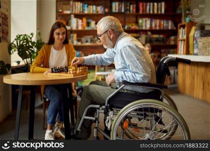 Disabled grandfather in wheelchair and granddaughter play chess, disability, cafeteria interior on background. Handicapped older male person and young female nurse, paralyzed people in public places. Disabled grandfather and granddaughter play chess