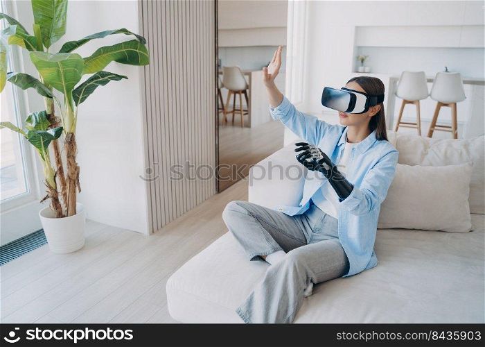 Disabled person gets rehabilitation and playing in cyber space. Young caucasian woman sitting in living room on couch. Handicapped girl in vr glasses. Amputee with bionic limb prosthesis.. Disabled person gets rehabilitation and playing in cyber space. Young caucasian woman in vr glasses.