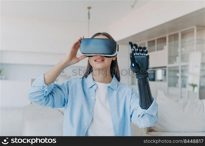 Disabled person gets rehabilitation. Handicapped girl with cyber arm in vr goggles at home. Young european woman sitting in living room on couch is excited. Lady touches the vision.. Disabled person gets rehabilitation. Handicapped girl with cyber arm in vr goggles at home.