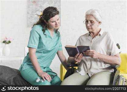 disabled senior female patient sitting wheel chair reading book with nurse