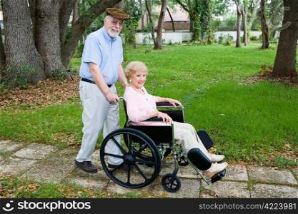 Disabled senior woman being pushed through the park in a wheelchair by her loving husband.