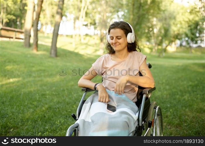 Disabled woman in wheelchair listens to music with headphones in park. Paralyzed people and disability, handicap overcoming. Handicapped female person walking in park. Disabled woman listens to music with headphones