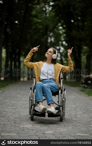 Disabled woman in wheelchair listens to music with headphones in summer park. Paralyzed people and disability, handicap overcoming. Handicapped female person in public place. Disabled woman in wheelchair listens to music