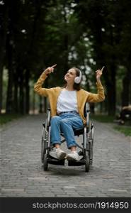 Disabled woman in wheelchair listens to music with headphones in summer park. Paralyzed people and disability, handicap overcoming. Handicapped female person in public place. Disabled woman in wheelchair listens to music