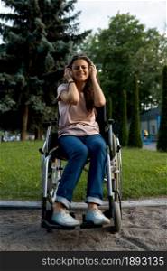 Disabled woman in wheelchair listens to music with headphones. Paralyzed people and disability, handicap overcoming. Handicapped female person walking in park. Disabled woman in wheelchair listens to music