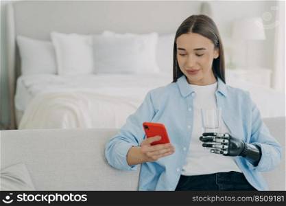 Disabled woman with phone. Girl is using futuristic robotic arm prosthesis and holding glass of water. European woman with artificial arm in morning in her bedroom. Amputee lifestyle after surgery.. Disabled woman with phone holding glass of water with robotic arm prosthesis in bedroom.