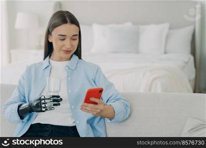 Disabled woman with phone. Girl is using futuristic robotic arm prosthesis and holding glass of water. European woman with artificial arm in morning in her bedroom. Amputee lifestyle after surgery.. Disabled woman with phone holding glass of water with robotic arm prosthesis in bedroom.