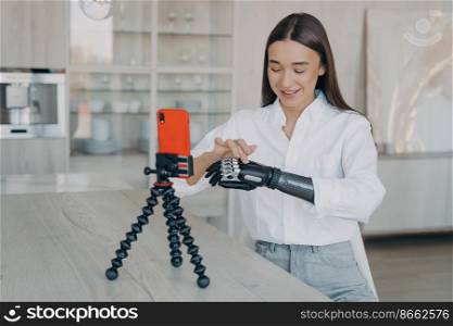 Disabled young girl blogger showing her artificial robotic hand her followers, using smartphone. Female talking about bionic prosthetic arm in live air in social networks, streaming from home.. Disabled young girl blogger showing her artificial robotic arm her followers, using smartphone