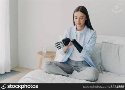 Disabled young girl disassembles bionic prosthetic arm, sitting on bed at home. Female with disability checking her artificial robotic limb in bedroom. Medical technologies concept.. Disabled young girl disassembles bionic prosthetic arm, sitting on bed at home. Medical technologies