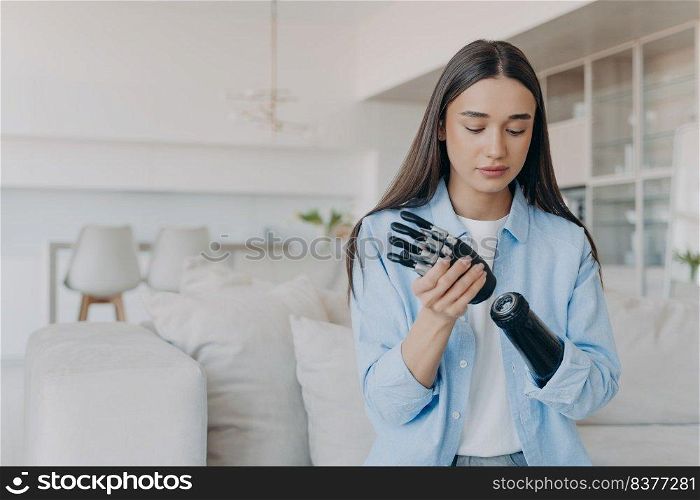 Disabled young woman getting used to new bionic prosthesis. Cyber sensor hand has processor chip and buttons. European girl setting her carbon robotic hand functions. Handicapped woman in living room.. Disabled young woman getting used to new bionic prosthesis. Girl setting her robotic hand functions.