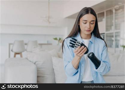 Disabled young woman getting used to new bionic prosthesis. Cyber sensor hand has processor chip and buttons. European girl setting her carbon robotic hand functions. Handicapped woman in living room.. Disabled young woman getting used to new bionic prosthesis. Girl setting her robotic hand functions.