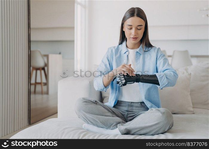 Disabled young woman is changing settings of bionic arm in morning. Cyber sensor hand has processor and buttons. European amputee adjusting electronic artificial arm at home. High tech carbon hand.. Disabled young woman is changing settings of bionic arm in morning. High tech carbon hand.