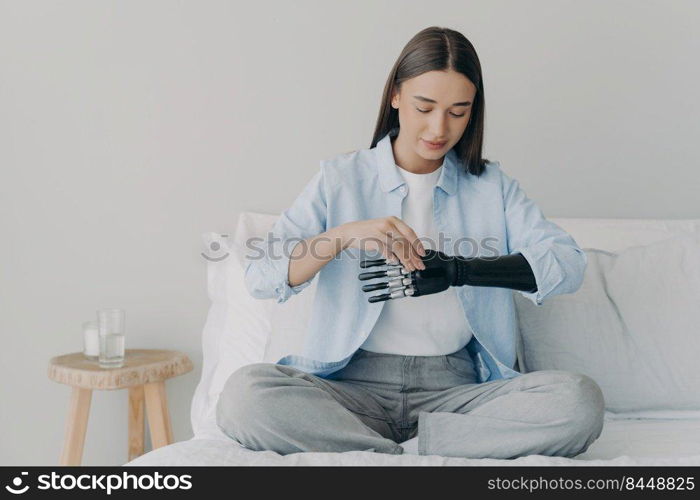 Disabled young woman is setting myoelectric prosthesis prosthesis parameters and functions. Bionic hand has processor chip and buttons. Attractive european girl adjusting her artificial arm at home.. Disabled young woman is setting myoelectric prosthesis prosthesis parameters and functions.