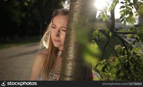 Disapointed woman leaning her head on tree trunk in park. Depressed girl with eyes full of sorrow, almost crying, standing alone near tree outdoors. Beautiful woman experiencing emotional stress and depression. Slow motion. Steadicam stabilized shot