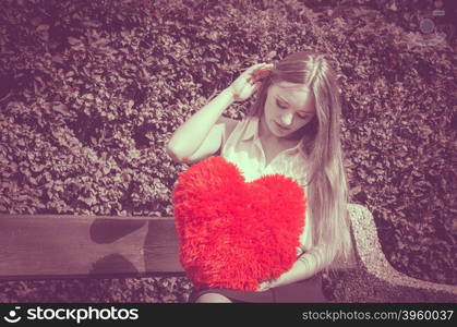 Disappointed woman with big red heart. Love and feelings. Unhappy hurt woman sitting on bench with big red heart in hands. Beautiful sad girl in park.