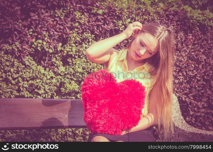 Disappointed woman with big red heart. Love and feelings. Unhappy hurt woman sitting on bench with big red heart in hands. Beautiful sad girl in park.