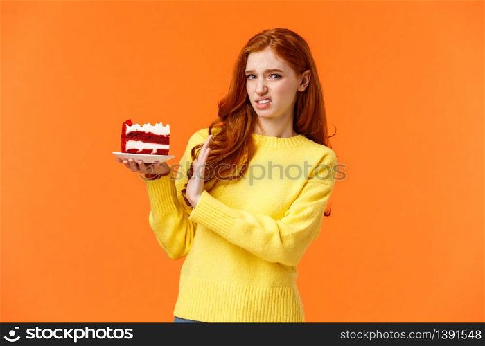 Disgusted and displeased, picky redhead girl look after body shape, dont eat sweets, holding piece tasty cake and refusing eat, showing stop sign, grimace at camera with aversion, orange background.. Disgusted and displeased, picky redhead girl look after body shape, dont eat sweets, holding piece tasty cake and refusing eat, showing stop sign, grimace at camera with aversion, orange background