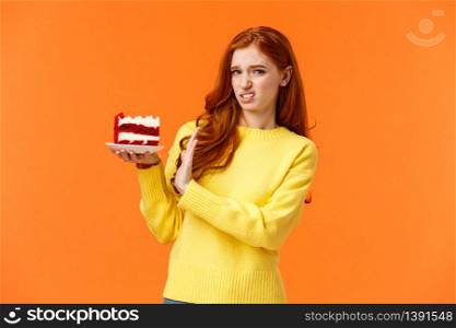 Disgusted and displeased, picky redhead girl look after body shape, dont eat sweets, holding piece tasty cake and refusing eat, showing stop sign, grimace at camera with aversion, orange background.. Disgusted and displeased, picky redhead girl look after body shape, dont eat sweets, holding piece tasty cake and refusing eat, showing stop sign, grimace at camera with aversion, orange background