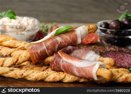 Dish for Antipasto snacks with salami, bread sticks (Grissini) wrapped with prosciutto olives and cheese paste on the wooden surface