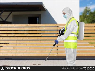 disinfection service and pandemic concept - sanitation worker in protective gear or hazmat suit, medical mask, gloves and goggles with pressure washer or sanitizer over house on city street background. sanitation worker in hazmat with pressure washer