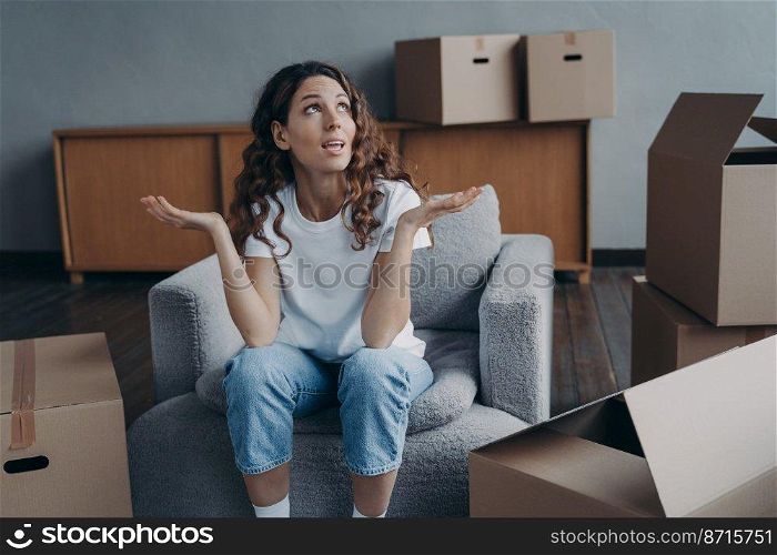 Displeased divorced young woman exclaiming shrugging sitting on armchair with cardboard boxes, tired after packing things for relocation. Divorce, eviction, hard moving day concept.. Puzzled divorced girl exclaims shrugging sitting with boxes for relocation. Divorce, hard moving day