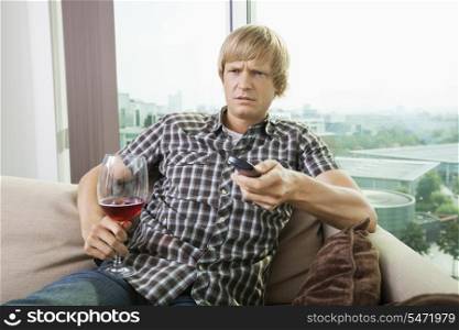 Displeased mid-adult man with wine glass watching television on sofa at home