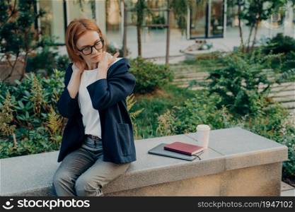 Displeased redhead European woman executive worker suffers from neck pain stiffness in shoulders outdoor in park with takeaway coffee notepad ad notebook wears formal clothes and spectacles.. Displeased redhead European woman executive worker suffers from neck pain stiffness in shoulders