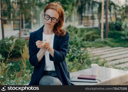 Displeased redhead woman suffers from pain in wrist after handwriting poses outdoor in urban park takes rest on bench wears formal clothes touches hand. Businesswoman feels tired after work.. Displeased redhead woman suffers from pain in wrist after handwriting poses outdoor