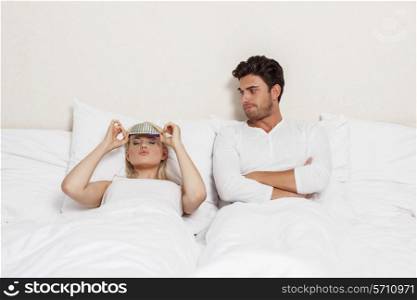 Displeased young man looking at woman wearing eye mask in bed
