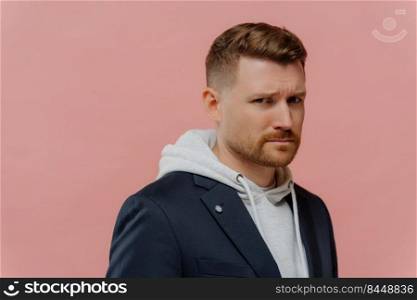 Displeased young man with bristle frowns face has offended look feels very upset dressed in casual hoodie and black jacket stands sideways against pink studio background copy space for your text