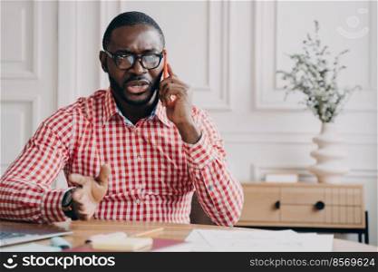 Dissatisfied stressed African American businessman emotionally speaking on mobile phone while sitting at workplace at home office, angry entrepreneur man arguing on telephone. Dissatisfied African American businessman emotionally speaking on smartphone while sitting at desk