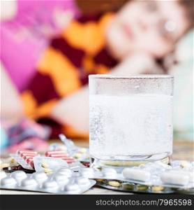 dissolving drug in glass with water and pills on table close up and sick woman with scarf around her neck on sofa in living room on background