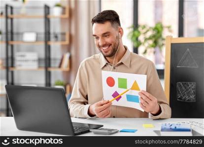 distance education, primary school and remote job concept - happy smiling male teacher with laptop and picture of geometric shapes in different colors having online class at home office. teacher showing shapes at online class at home