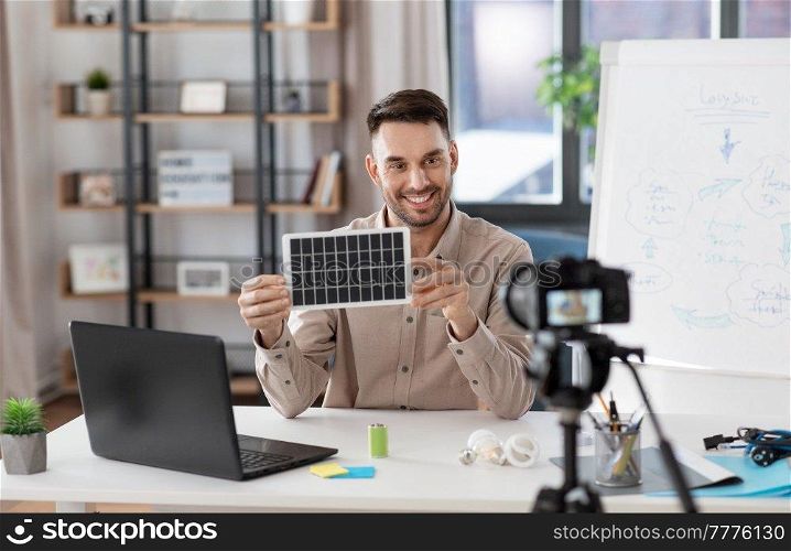 distance education, school and green energy concept - happy smiling male teacher with camera and solar battery model having online class at home office. male teacher with camera and solar battery at home