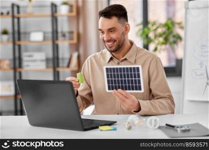 distance education, school and green energy concept - happy smiling male teacher with laptop computer and solar battery model having online class at home office. male teacher with laptop and solar battery at home