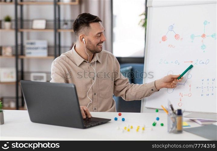 distance education, school and remote job concept - happy smiling male chemistry teacher in earphones with laptop computer having online class and showing molecular model on flip chart at home office. chemistry teacher with laptop having online class