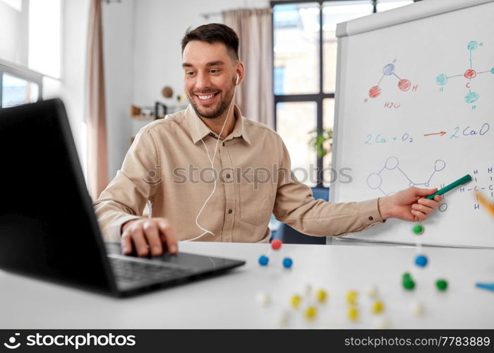 distance education, school and remote job concept - happy smiling male chemistry teacher with laptop computer having online class and showing chemical formula on flip chart at home office. chemistry teacher with laptop having online class