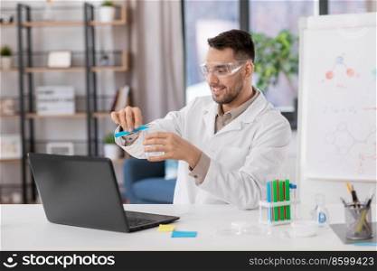 distance education, school and science concept - happy smiling male chemistry teacher in goggles with laptop computer having online class and pouring chemical from test tube to flask at home office. chemistry teacher with laptop having online class