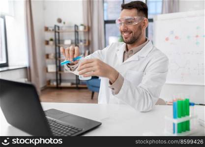 distance education, school and science concept - happy smiling male chemistry teacher in goggles with laptop computer having online class and pouring chemical from test tube to flask at home office. chemistry teacher with laptop having online class