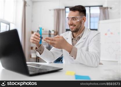 distance education, school and science concept - smiling male chemistry teacher in goggles and earphones with laptop computer having online class and showing test tube with chemical at home office. chemistry teacher with laptop having online class