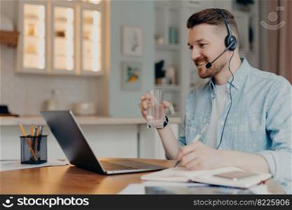 Distance learning concept. Millennial guy student in headset watches webinar has online lessons during quarantine enjoys internet education drinks water from glass makes notes poses at desktop