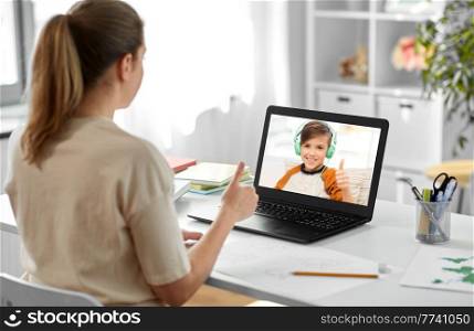 distant education, remote job and e-learning concept - female teacher with laptop computer having video call or online class with smiling student boy in headphones at home office and showing thumbs up. teacher with laptop having online class at home