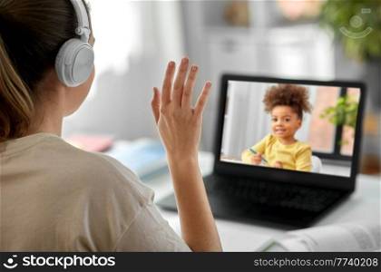 distant education, remote job and people concept - female teacher in headphones with laptop computer having video call or online class with little student girl at home and waving hand. teacher with laptop having video call with girl