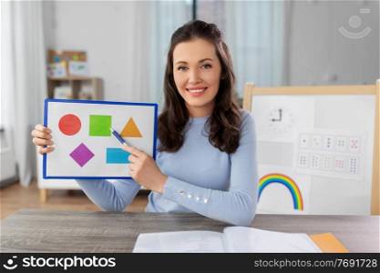 distant education, school and people concept - happy smiling female teacher with picture of geometric shapes in different colors having online class at home. teacher showing shapes in online class at home