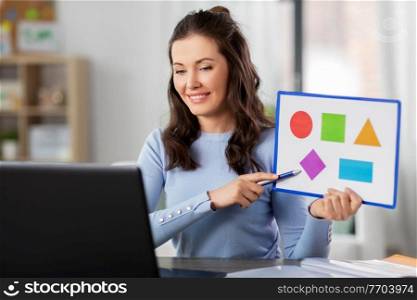distant education, school and people concept - happy smiling female teacher with laptop computer and picture of geometric shapes in different colors having online class at home. teacher showing shapes in online class at home