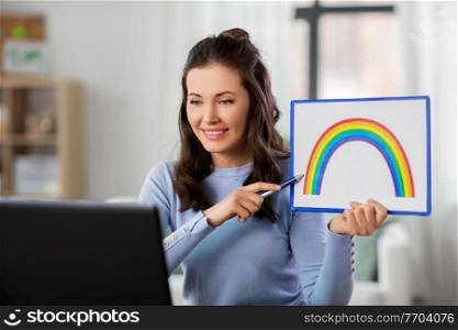 distant education, school and people concept - happy smiling female teacher with laptop computer and picture of rainbow having online art class at home. teacher with laptop having online class at home