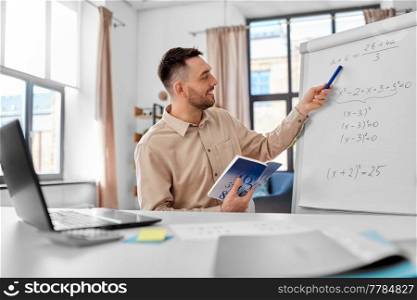 distant education, school and remote job concept - happy smiling male math teacher with laptop compute and book showing equation on flip chart having online class or video call at home office. math teacher with laptop has online class at home