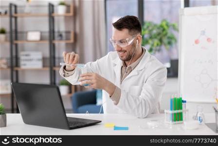 distant education, school and science concept - happy male chemistry teacher in goggles and earphones with laptop having online class and pouring chemical from test tube to flask at home office. chemistry teacher with laptop having online class
