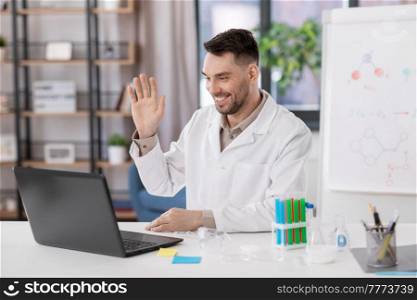 distant education, school and science concept - happy smiling male chemistry teacher with laptop computer having online class and waving hand at home office. chemistry teacher with laptop having online class